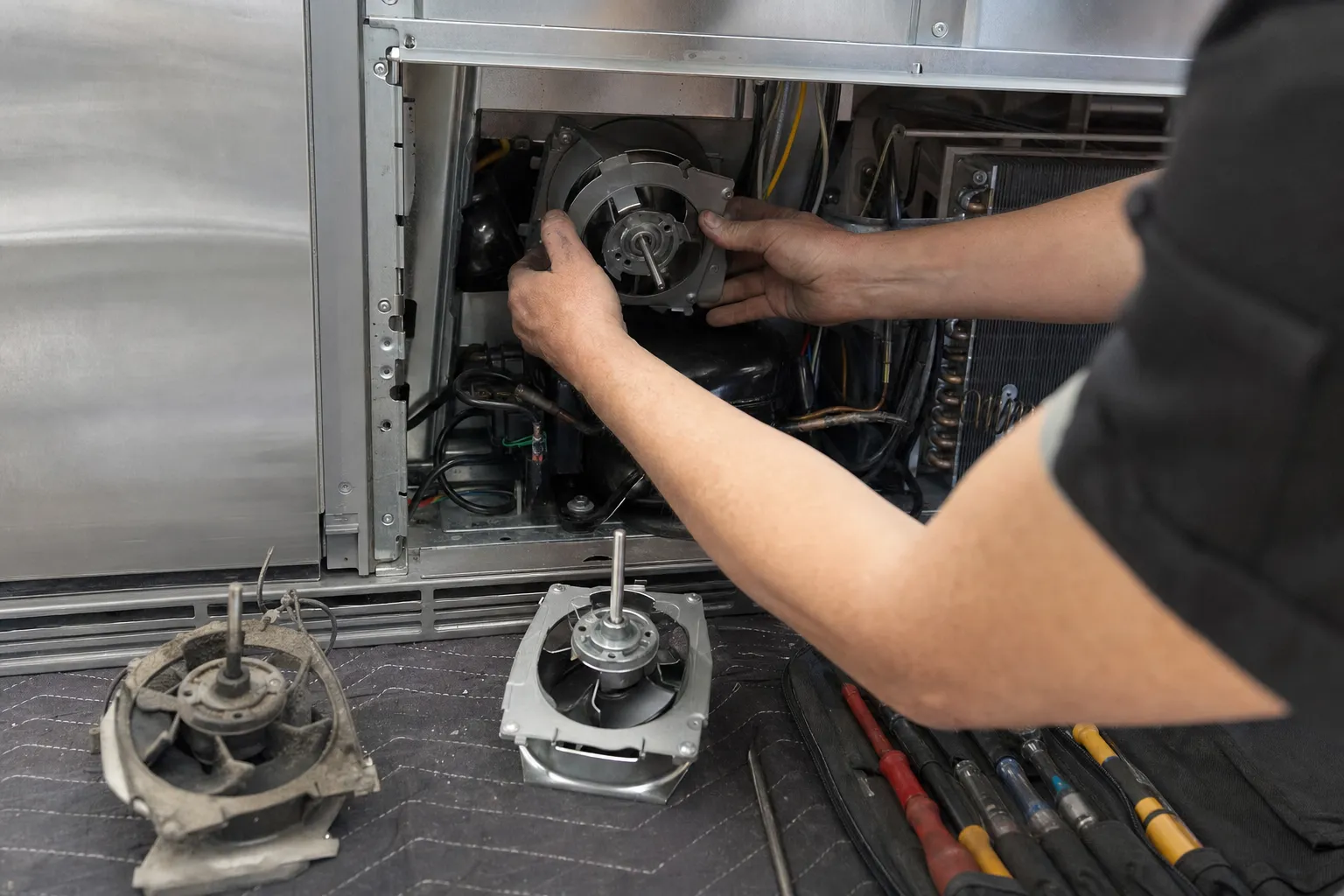 Fan motor being seated in the machine compartment of a stainless built-in, two removed fan assemblies and a tool roll on the mat below