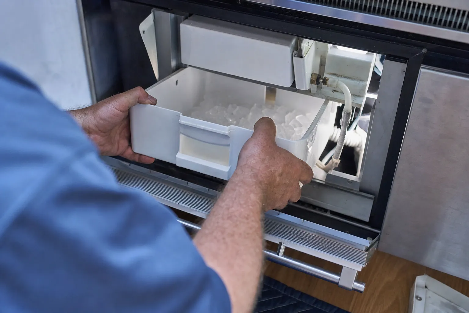 Bin of ice cubes being lifted out below the white ice-maker housing of a stainless unit, a brass fitting and white water tube down one side